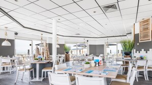 Un comedor amplio con mesas y sillas blancas, una isla central con una planta, y vista al mar a través de grandes ventanales.