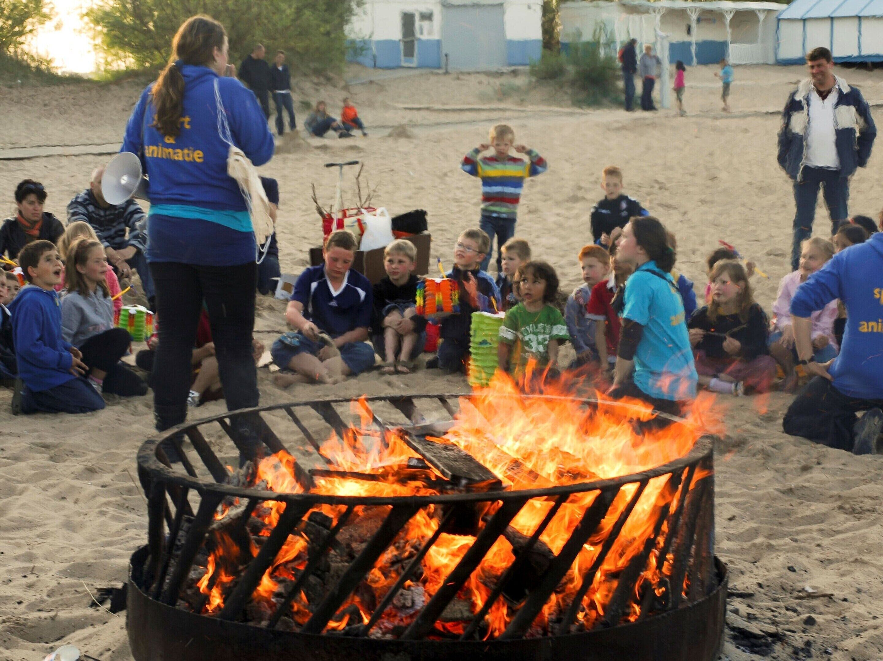 A group of children gathered around a campfire on a sandy beach, with adults supervising.