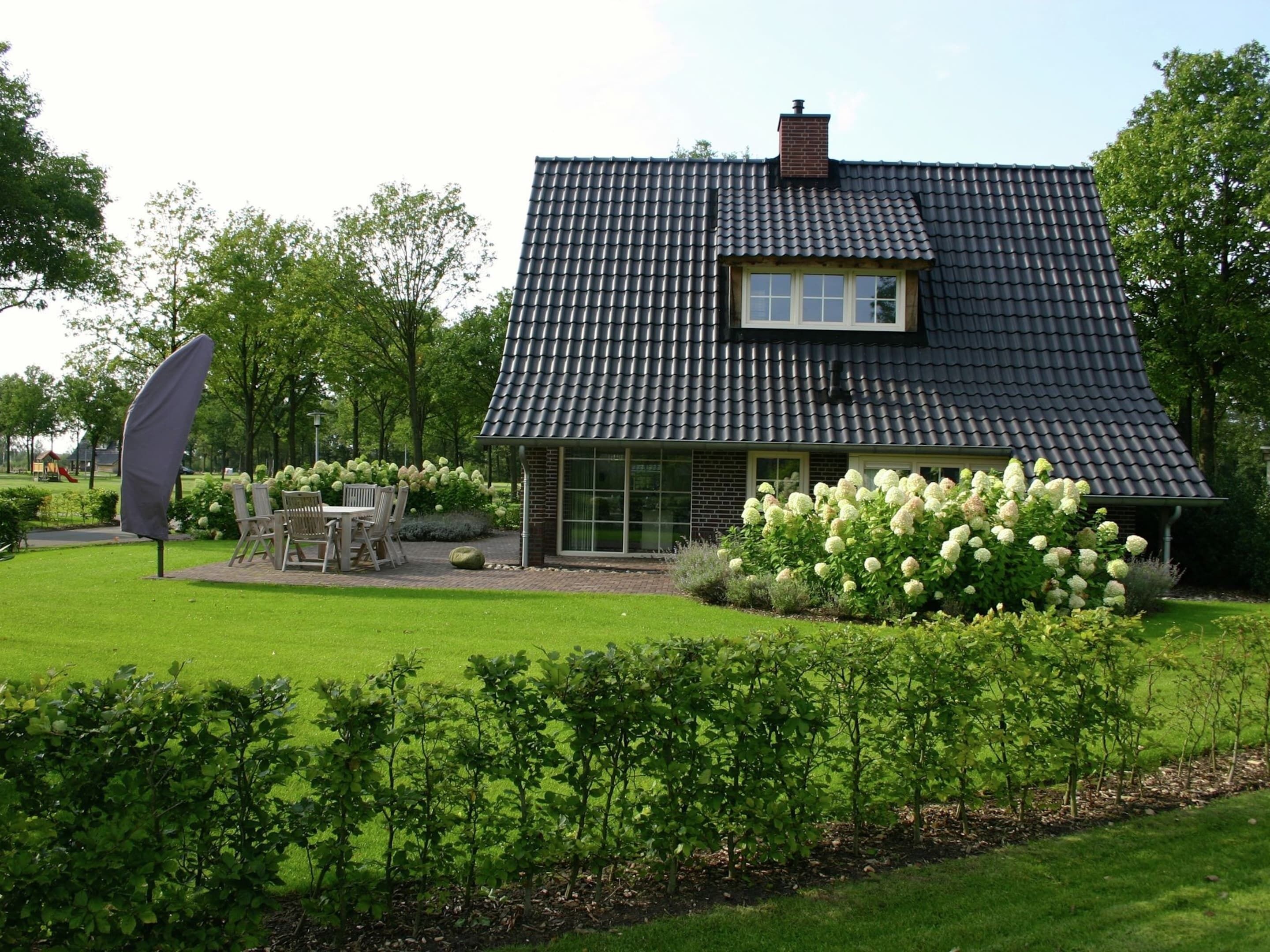 Ett hus med ett mörkt tak täckt med takpannor, en tegelmurad skorsten och ett stort fönster. Det finns en uteplats med utemöbler och en trädgård med vita blommor.