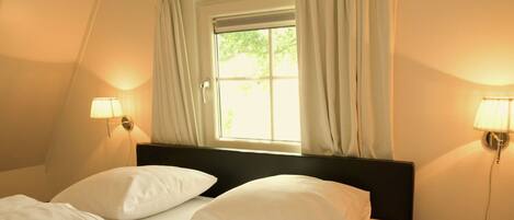 A hotel room with a bed, two bedside lamps, a window with curtains, and a nightstand with a magazine.