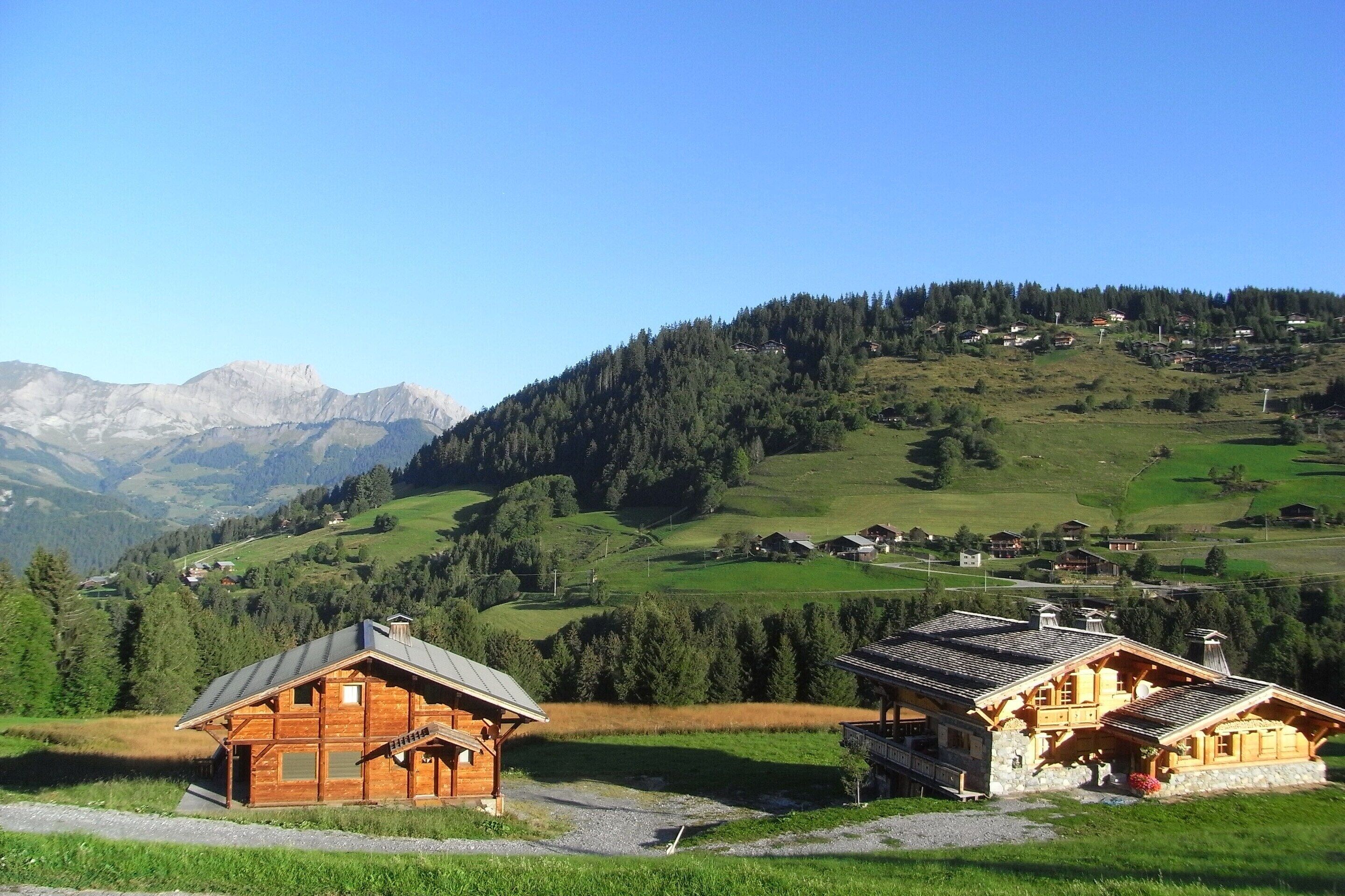 A wooden cabin with a sloped roof, surrounded by a lush green landscape and distant mountains.