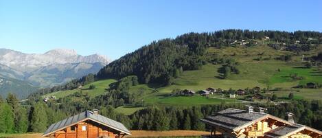 A wooden cabin with a sloped roof, surrounded by a lush green landscape and distant mountains.