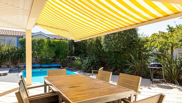 Une terrasse extérieure avec une table et des chaises en bois, un auvent à rayures jaunes, une piscine et un barbecue.