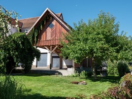 Una casa de madera tradicional con techo inclinado, un porche de madera y un jardín bien cuidado con césped y arbustos.