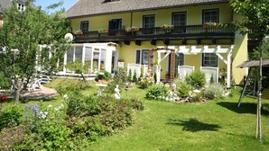 A two-story building with a balcony, a garden with various plants and statues, and a clear blue sky. - Apartment in Feld am See by Brennsee Lake (Feld am See)