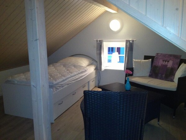 A small, cozy room with a sloped ceiling, a single bed, a small table, a chair, and a window with curtains. - Holiday Home by the Sea With Sauna (Sierksdorf)