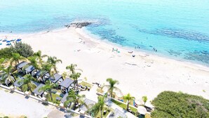 Vista aérea de una playa con agua azul cristalina, una orilla arenosa y una zona turística con sillas de descanso y sombrillas.