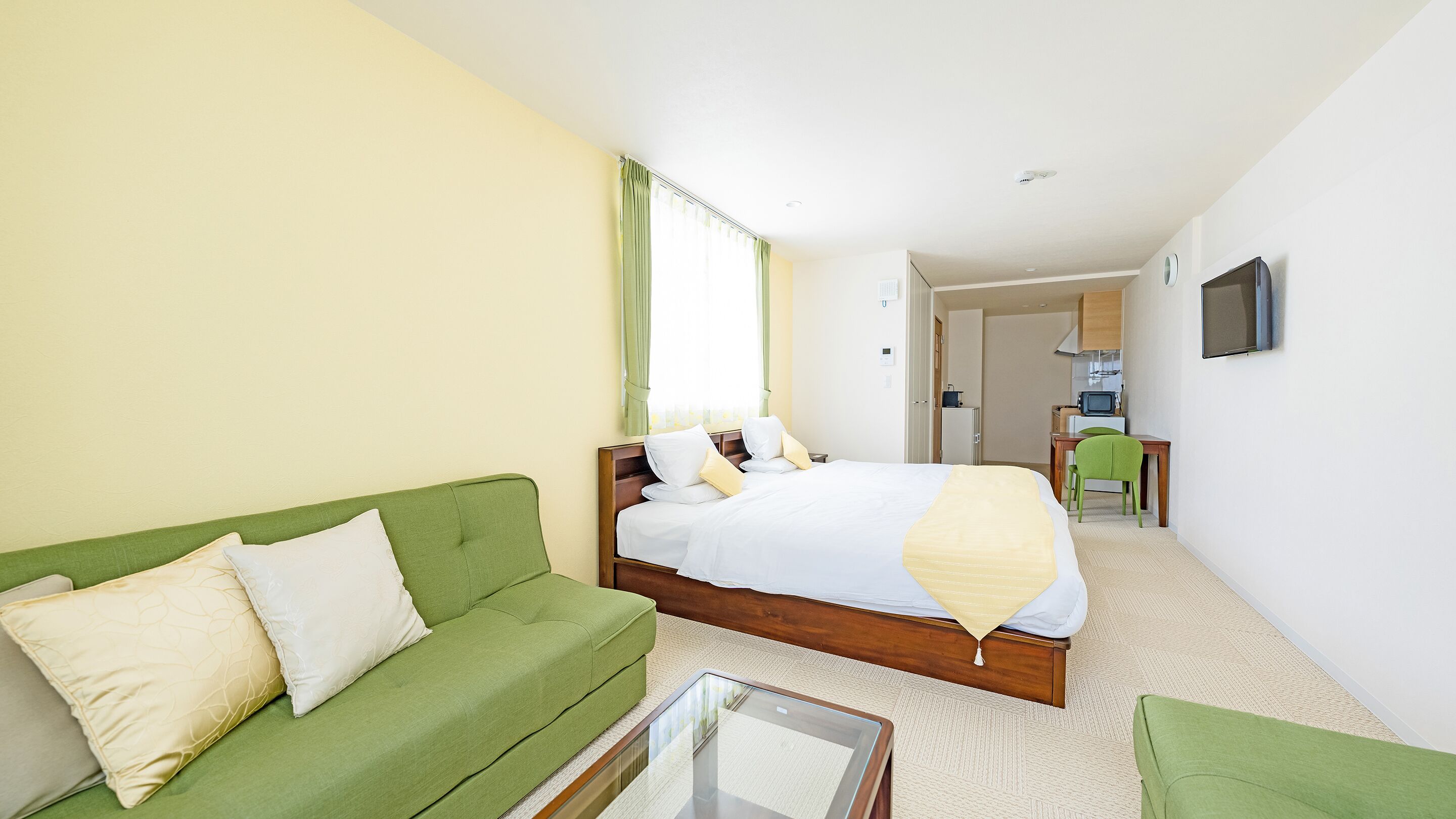 A hotel room with a green sofa, a bed with white bedding, a small table, and a kitchenette in the background.