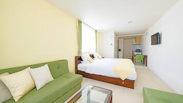 A hotel room with a green sofa, a bed with white bedding, a small table, and a kitchenette in the background.