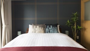 Una cama bien hecha con una colcha blanca y roja, dos almohadas con diseños abstractos, una planta alta en maceta y una pared oscura con un patrón de rejilla.