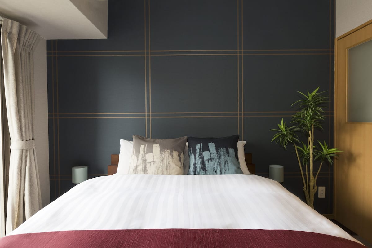 A neatly made bed with a white and red bedspread, two pillows with abstract designs, a tall potted plant, and a dark wall with a grid pattern.