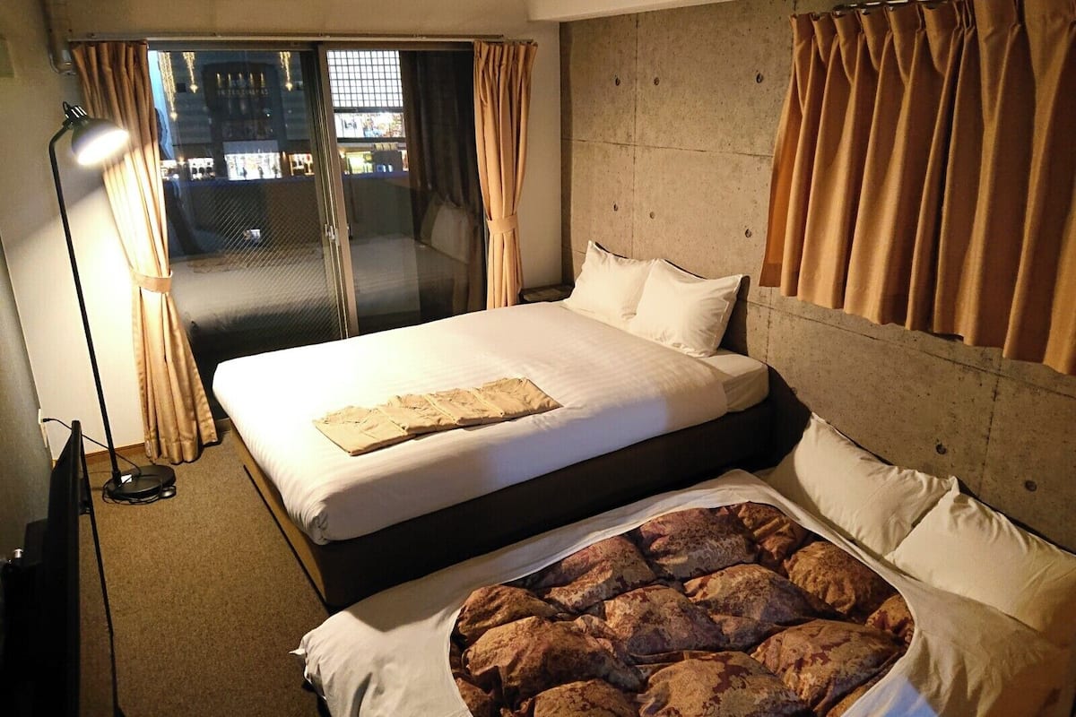 A hotel room with a large bed, a second smaller bed, a desk, and a view of the outside through a sliding door.