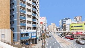 Un edificio de varios pisos con fachada azul y blanca, un letrero con caracteres japoneses y una calle concurrida con vehículos y una acera.