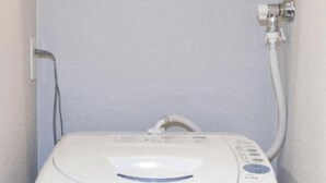 Calentador de agua portátil blanco y compacto, con panel de control, manguera y cable de alimentación, instalado en un baño pequeño con pared gris claro.