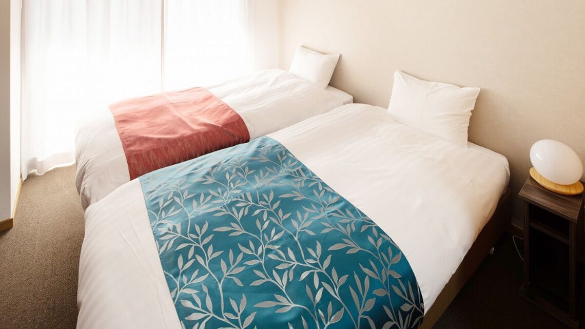 A hotel room with two beds, a bedside table with a lamp, and a window with curtains.