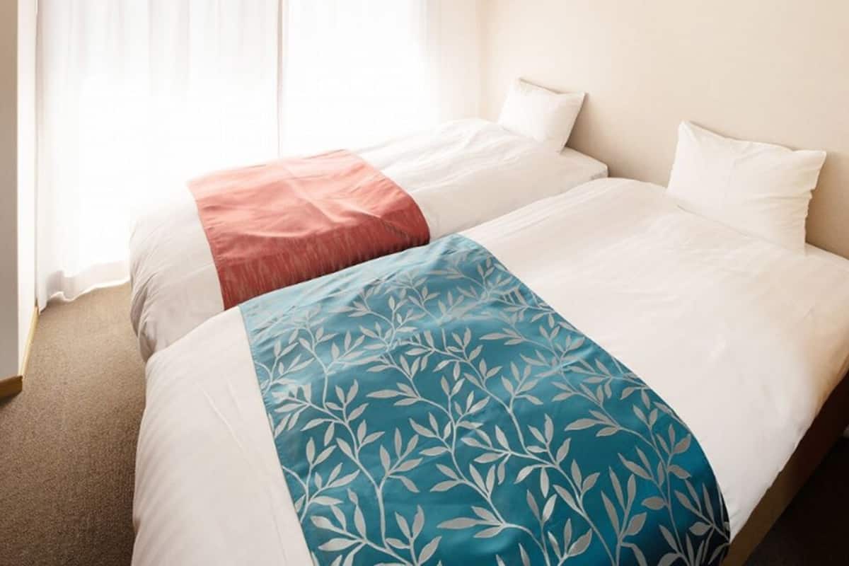 A hotel room with two beds, a bedside table with a lamp, and a window with curtains.