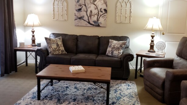 A living room with a dark brown leather sofa, a wooden coffee table, and two side tables with lamps. There is a large abstract painting on the wall and a patterned rug on the floor.