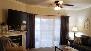 Una sala de estar con chimenea, televisor, ventilador de techo y balcón con cortinas.