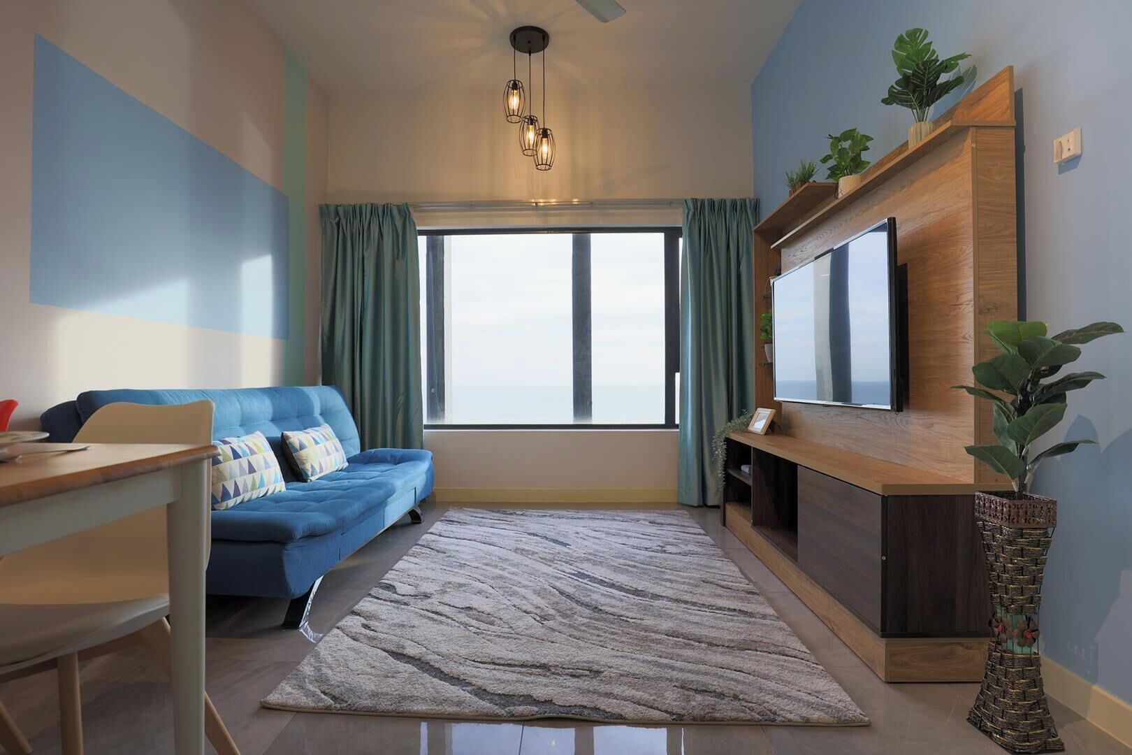 A modern living room with a blue sofa, a wooden TV stand, a large window with curtains, and a patterned rug.