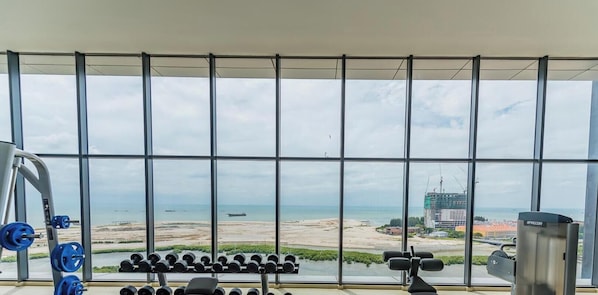 A gym with a view of a beach and a construction site through large windows. - Imperio Seaview Melaka By I Housing (Malacca City)