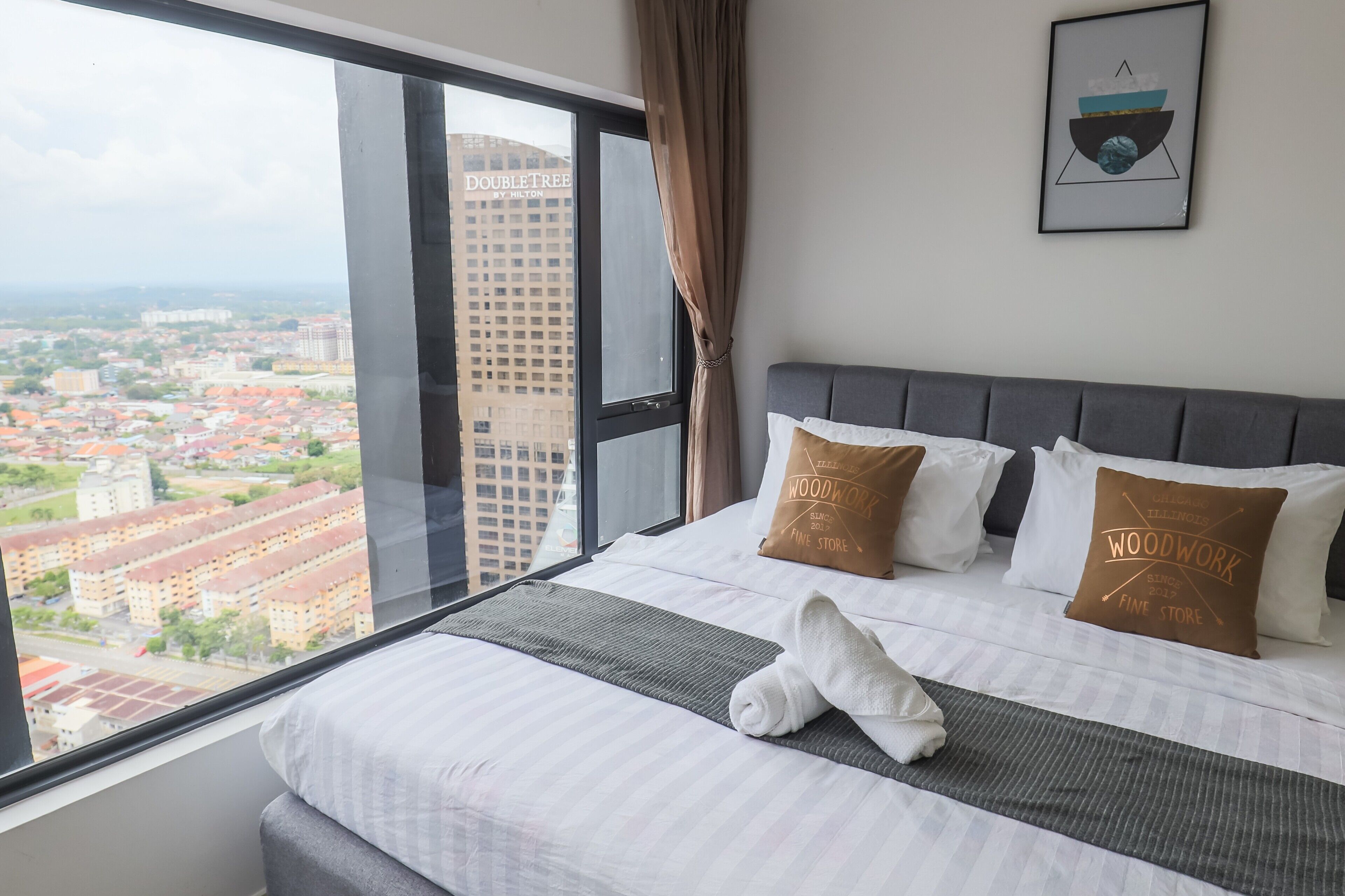 A hotel room with a large window, a bed with white and brown pillows, and a framed picture on the wall.