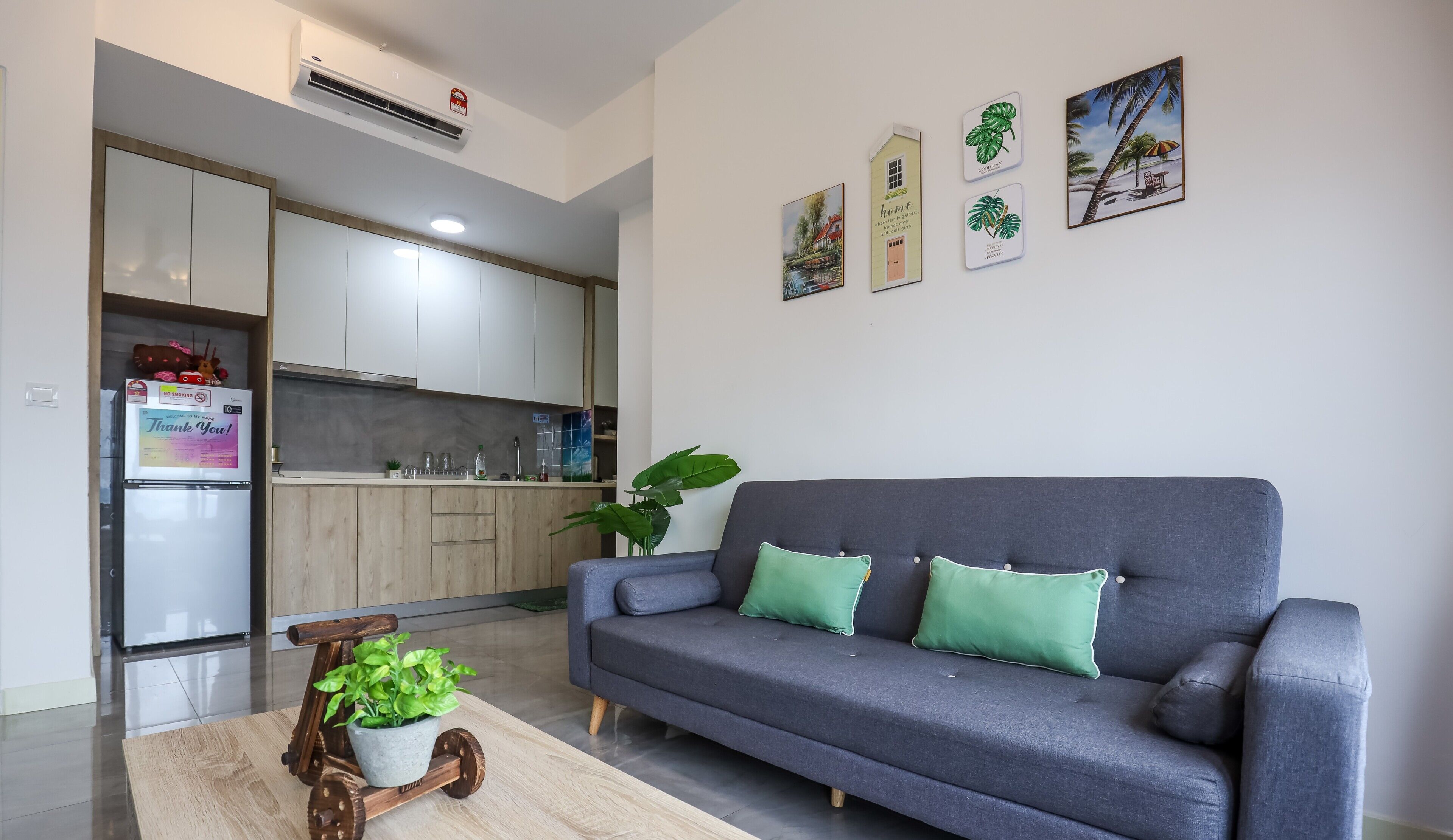 Een moderne woonkamer met een grijze bank, een houten salontafel met een plant, en een keuken op de achtergrond.
