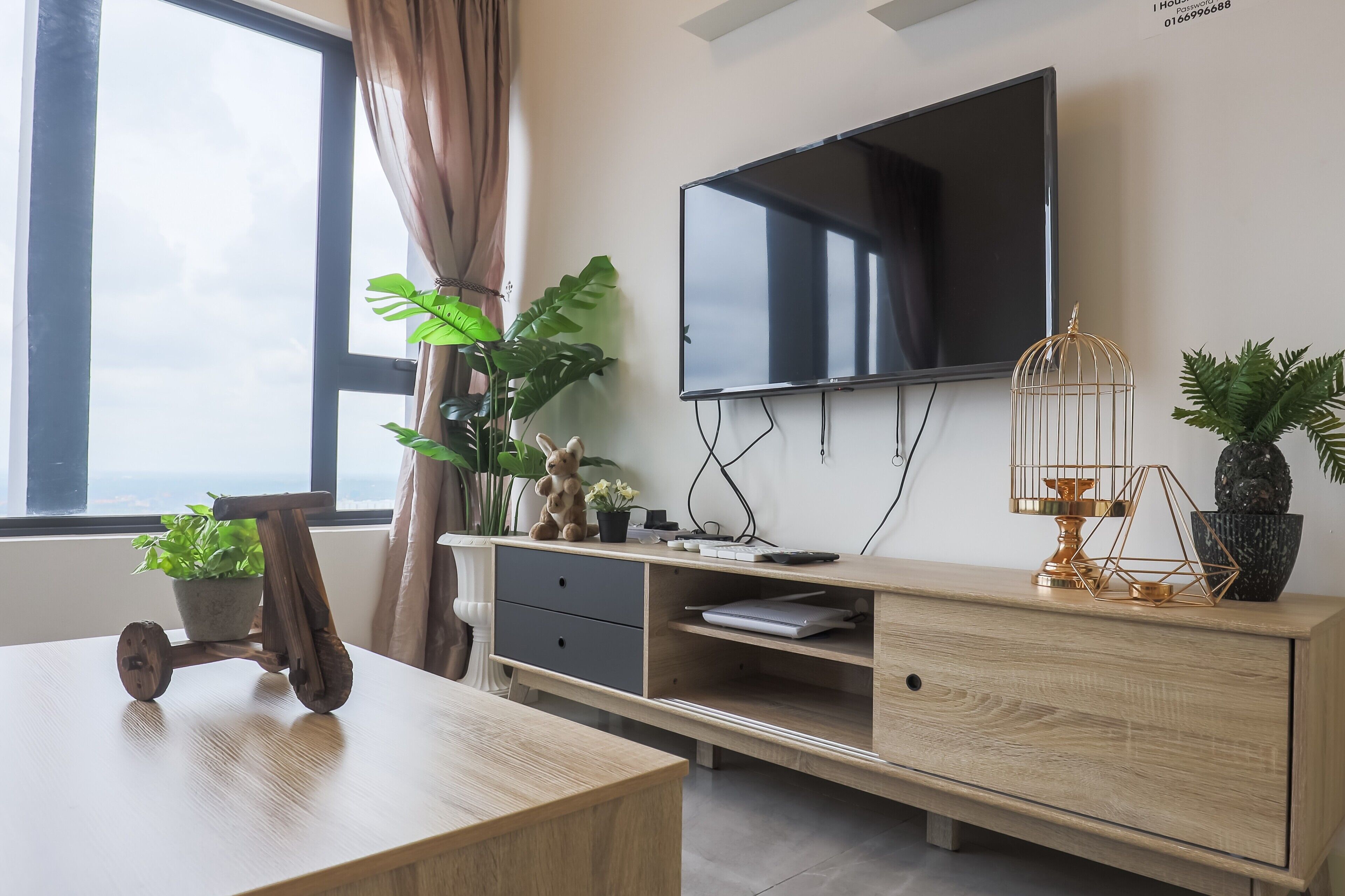 A modern living room with a flat-screen TV mounted on the wall, a wooden coffee table, a desk with a laptop, and various decorative items including plants and a birdcage.
