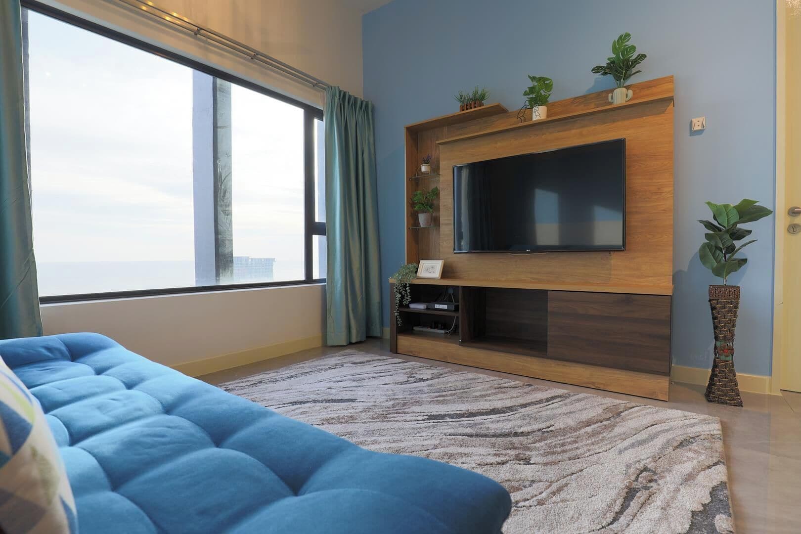 A living room with a large window, a blue sofa, a flat-screen TV mounted on a wooden wall, and a potted plant.