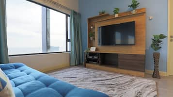 A living room with a large window, a blue sofa, a flat-screen TV mounted on a wooden wall, and a potted plant.