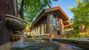 A rustic building with a stone fountain, large windows, and a wooden deck with chairs.