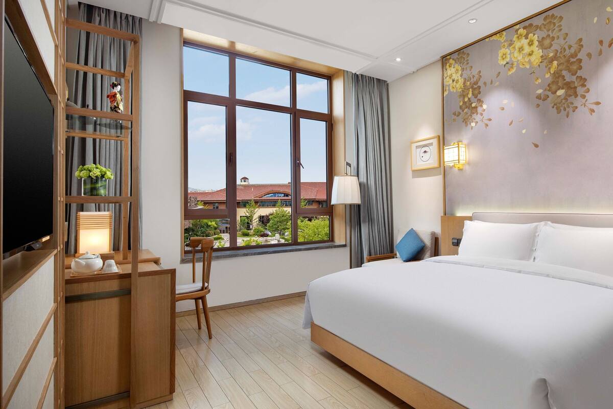 A hotel room with a large bed, a desk with a chair, a TV, and a window with a view of buildings and greenery.