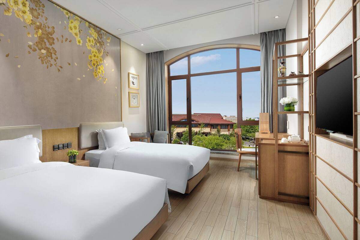 A hotel room with two beds, a large window with a view, a TV, and a desk with a chair.