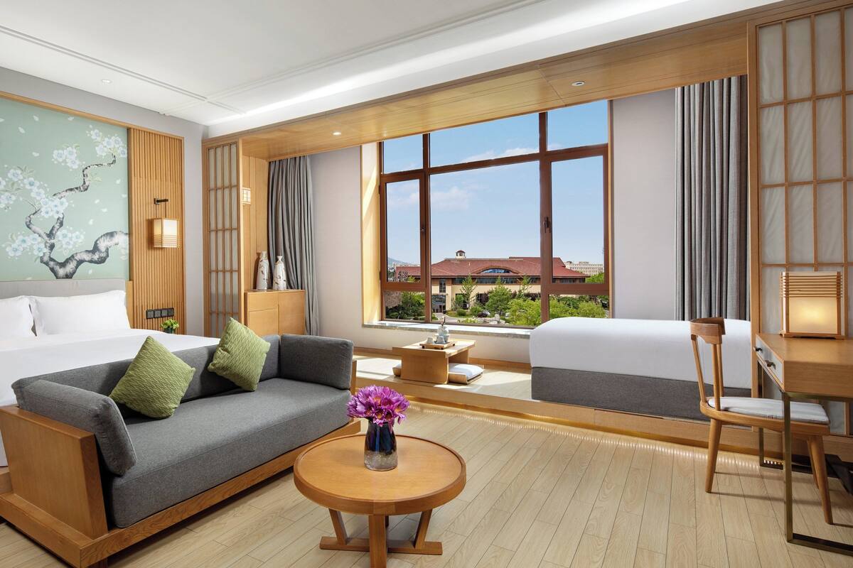 A hotel room with a bed, sofa, armchair, round wooden table, and a large window with a view of buildings.