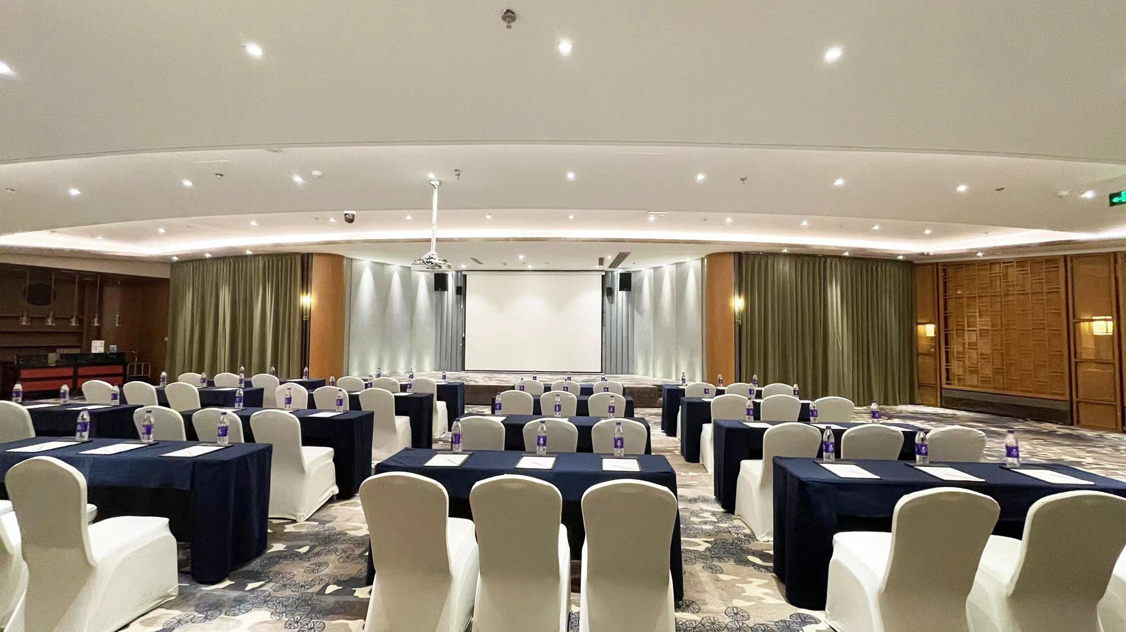 A conference room with rows of tables and chairs, a projector screen, and a stage.