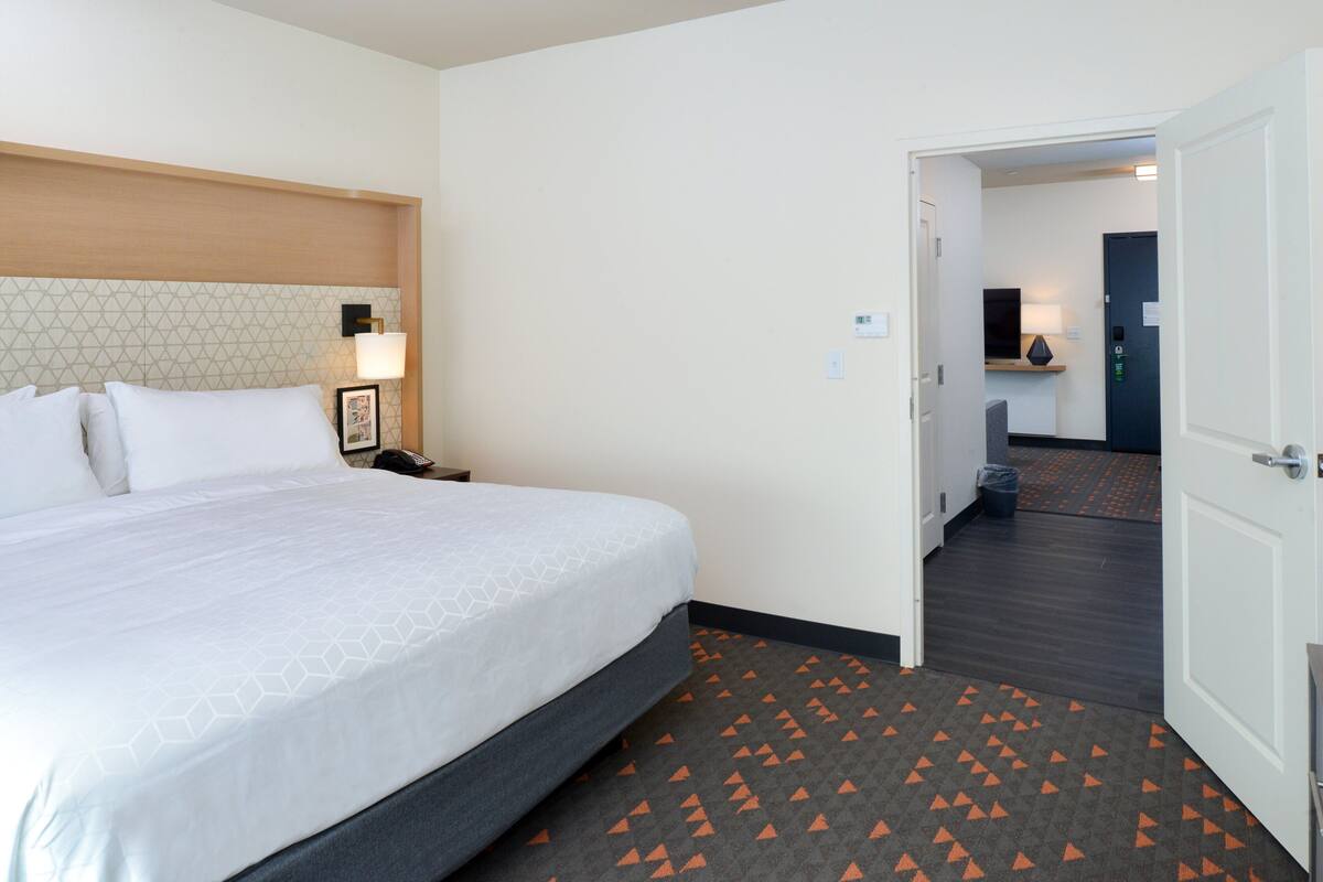 A hotel room with a large bed, a nightstand, a lamp, and a door leading to a hallway with a trash bin and a television.