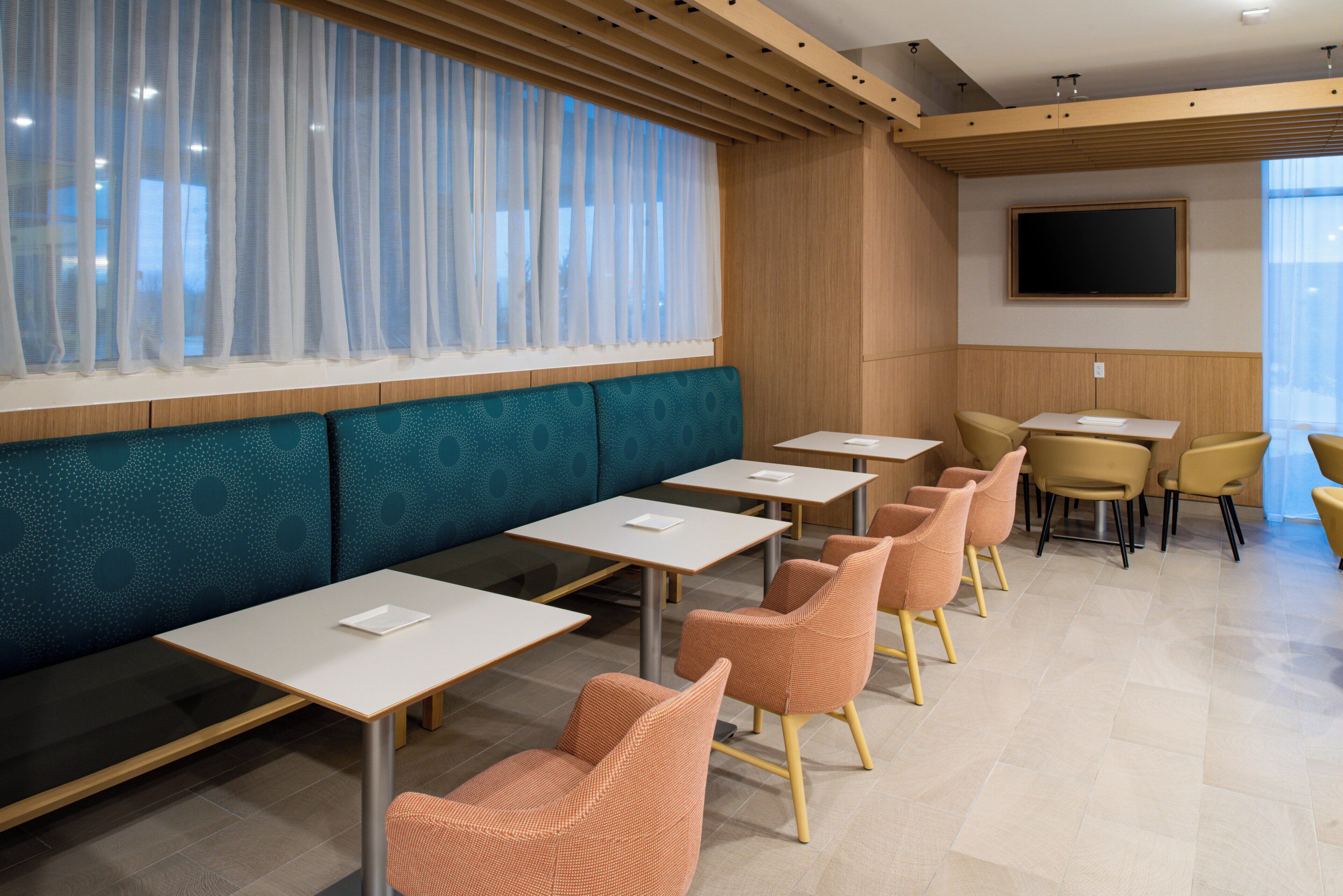 A modern dining area with booths and tables, a flat-screen TV on the wall, and a large window with sheer curtains.