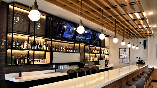 Zona de bar con una barra alargada, varios taburetes altos, lámparas colgantes y estantes llenos de botellas y vasos.