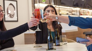 Un grupo de personas brindando con bebidas en un bar con decoración moderna y botellas de vino a la vista.