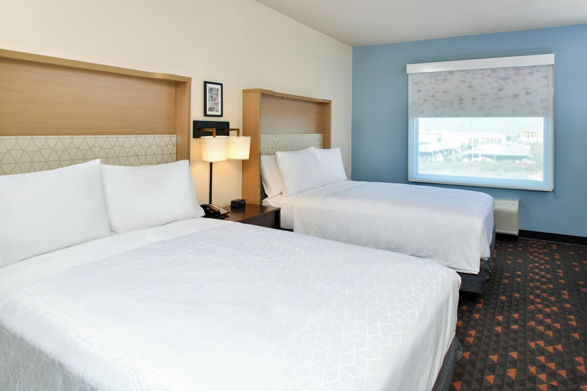 A hotel room with two beds, a nightstand, a lamp, a framed picture, and a window with a view.