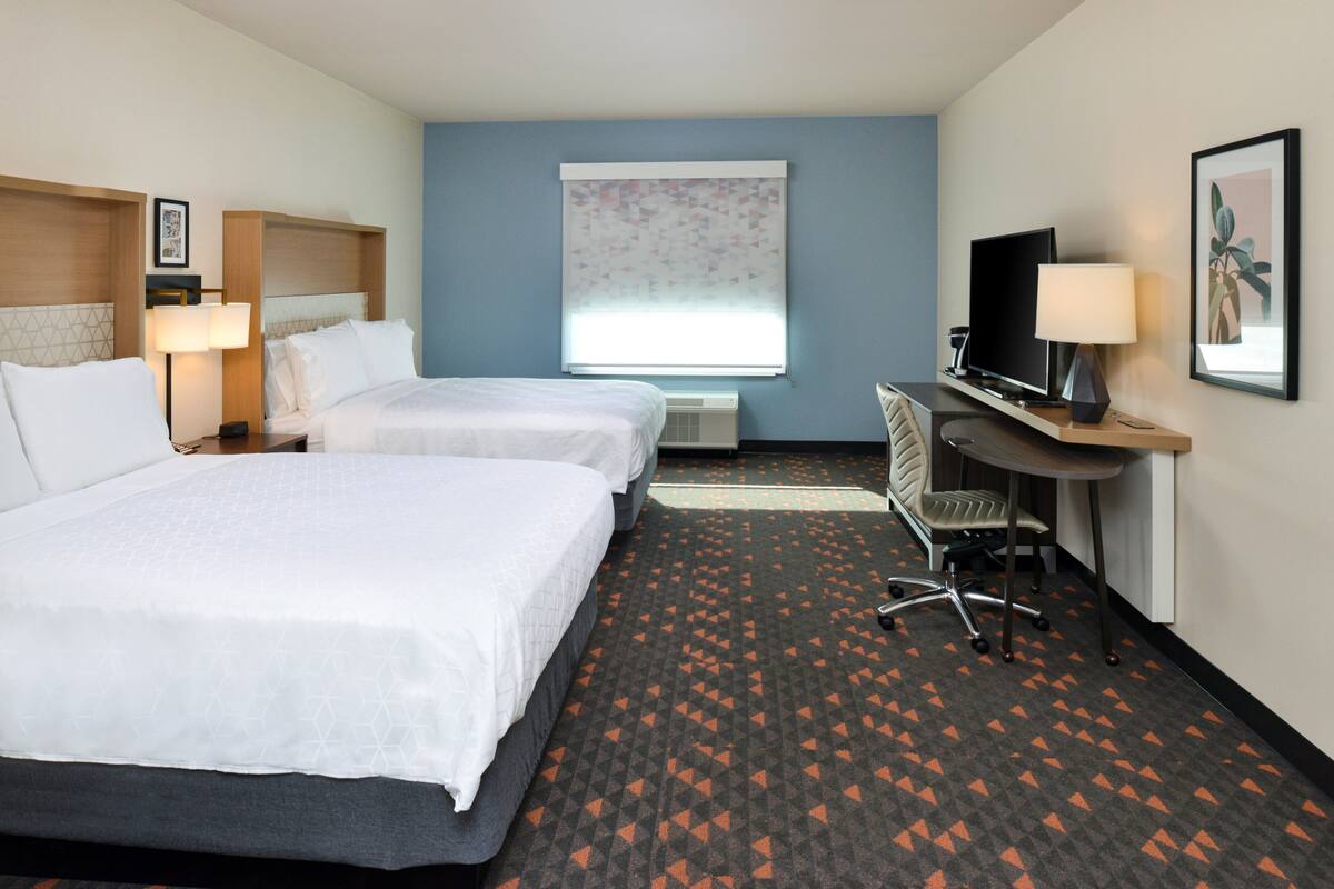 A hotel room with two beds, a desk with a television, a chair, and a window with blinds.