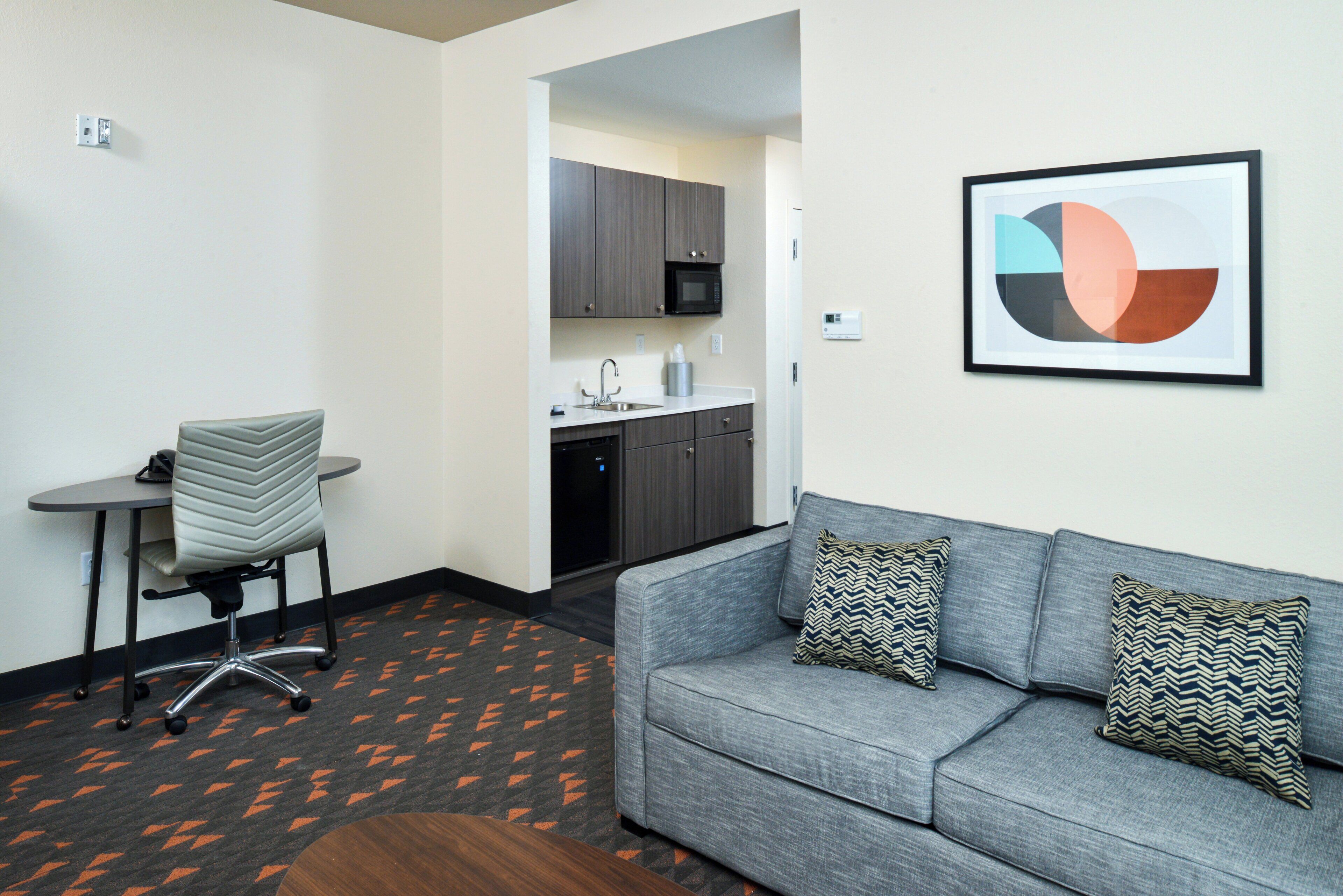 A modern hotel room with a gray sofa, patterned cushions, a small desk with a chair, a microwave, and a sink in the kitchenette.