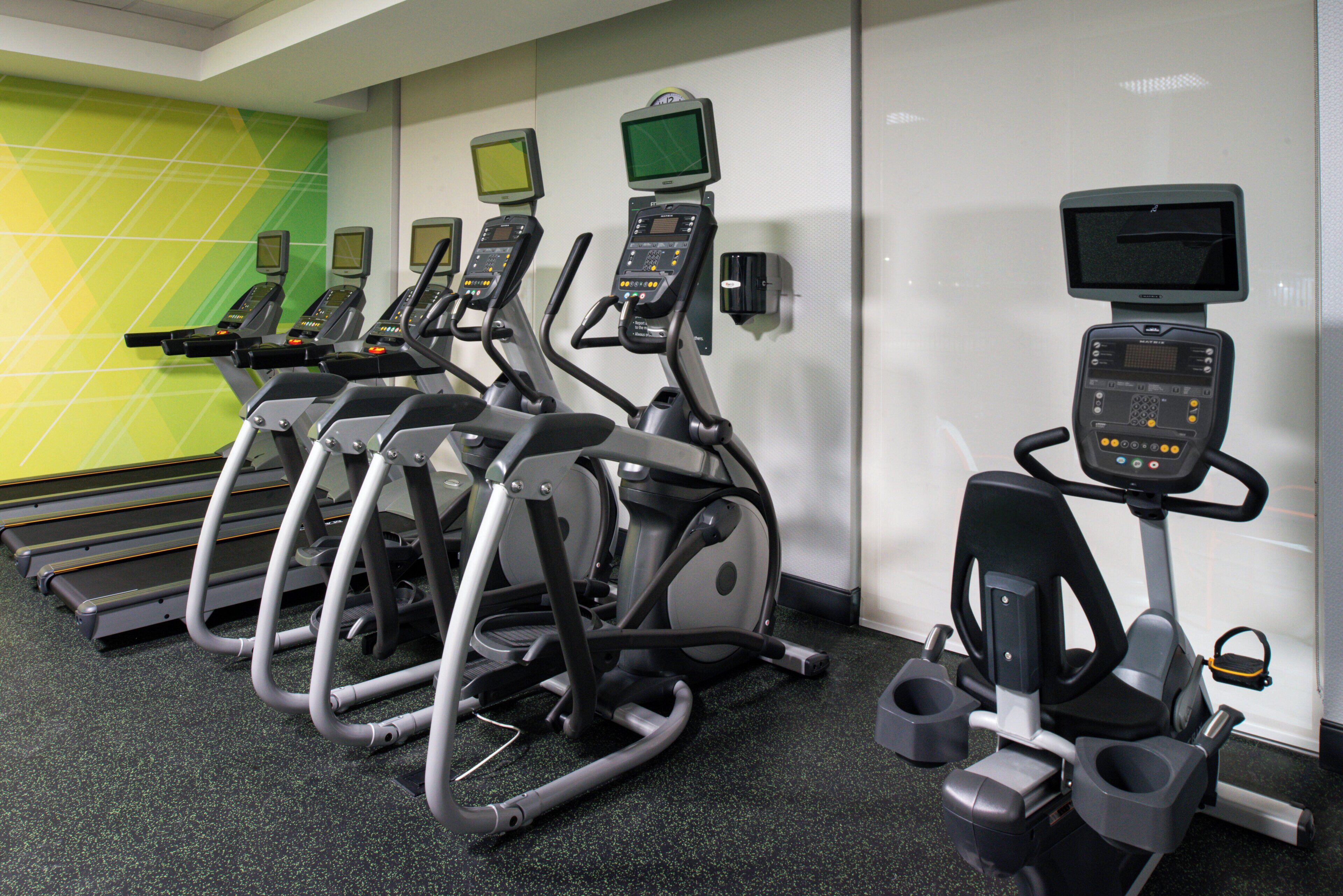 A row of stationary bikes with digital displays, arranged in a gym with a green and white tiled wall.