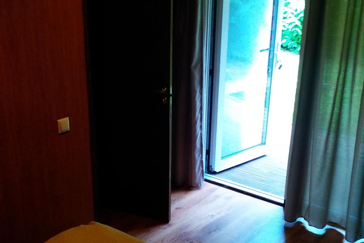 A room with a bed, a door leading to a balcony, and a view of greenery outside.
