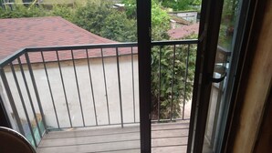 A balcony with a metal railing, a sliding glass door, and a view of a red-roofed building and greenery. - ATRIUM Guesthouse (Riga)