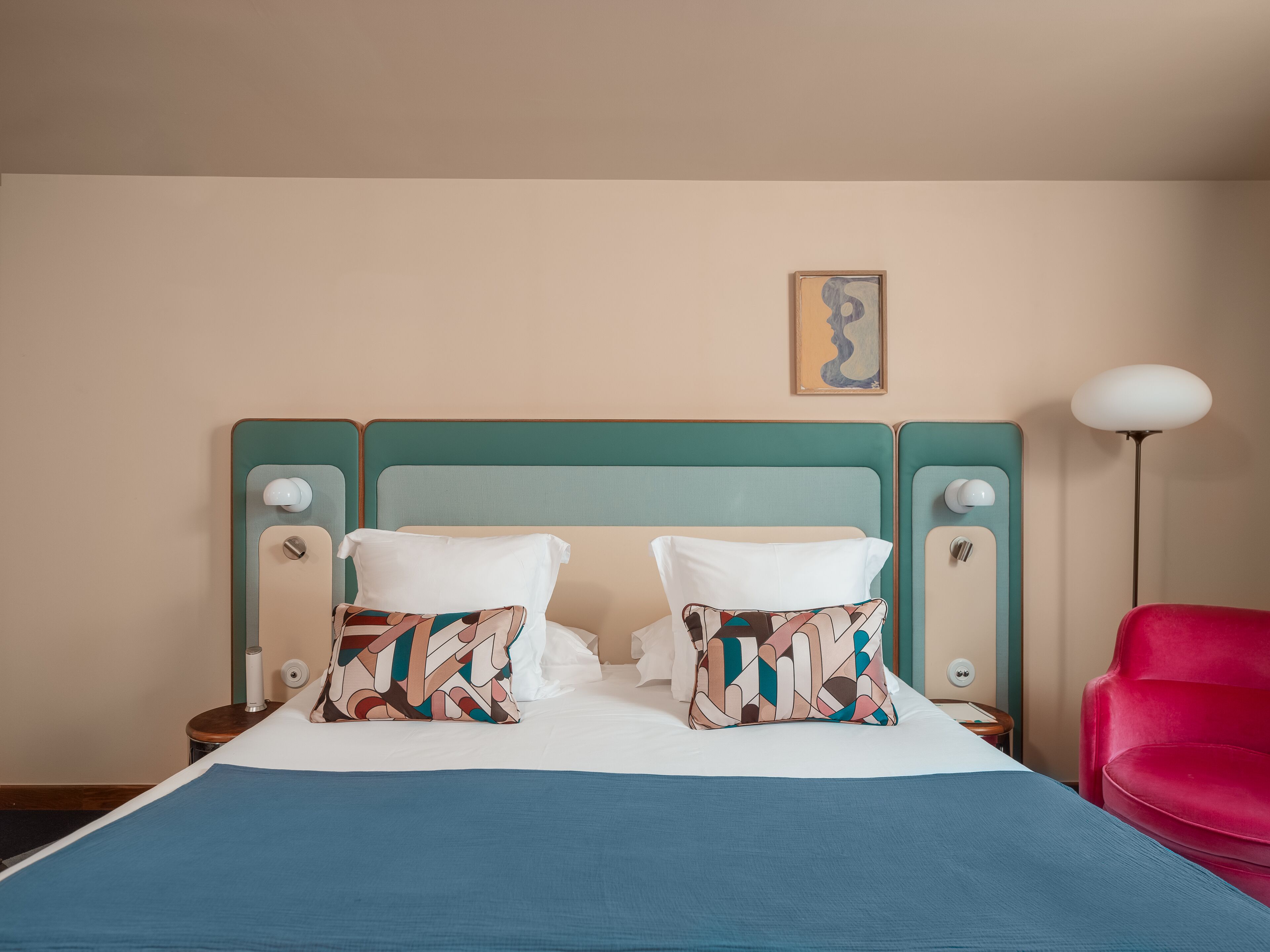 A bed with a teal headboard, two patterned pillows, a blue bedspread, and a red chair.