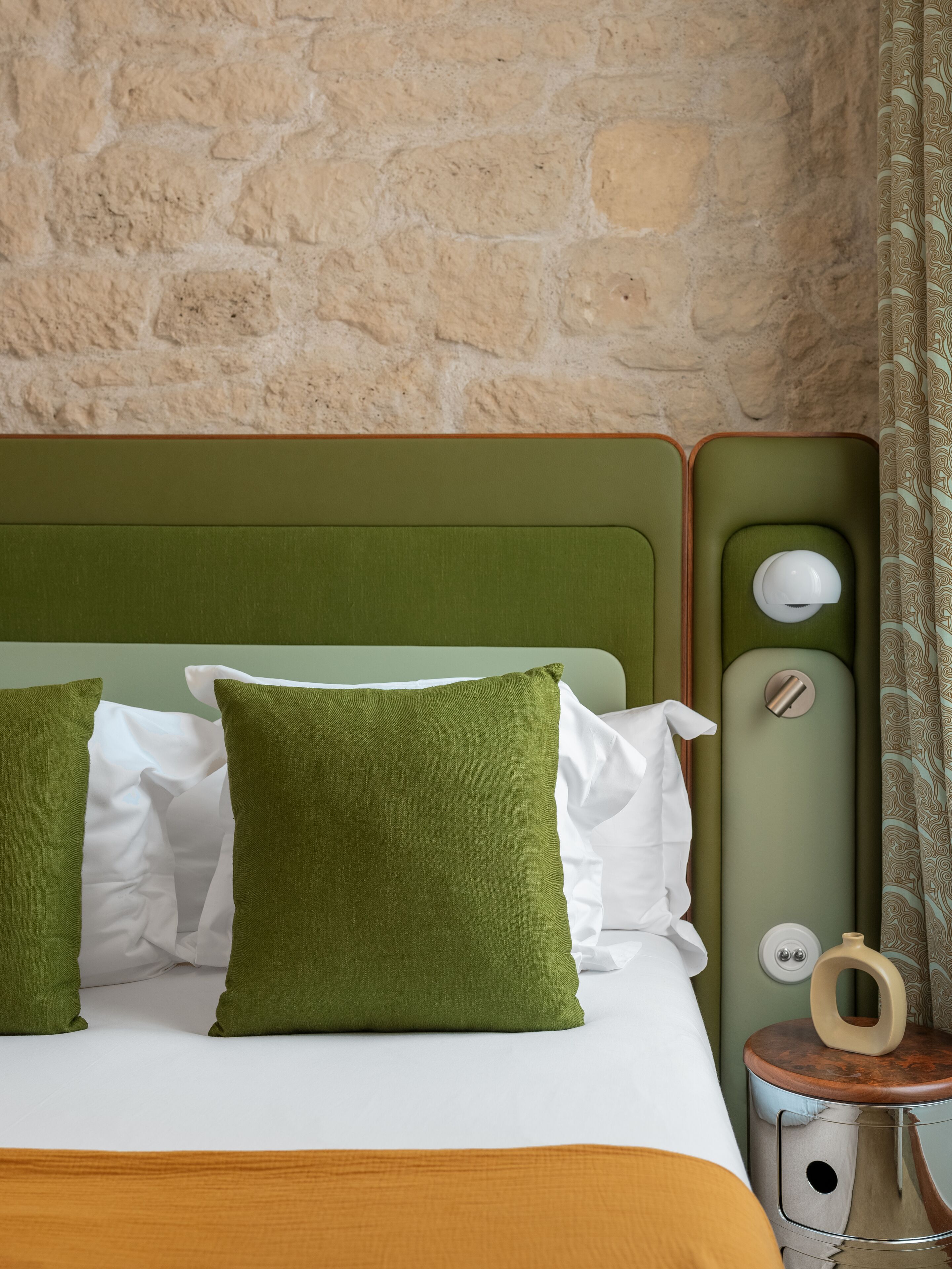 Un letto con cuscini verde e bianco, un comodino in legno con una lampada e una parete in pietra.