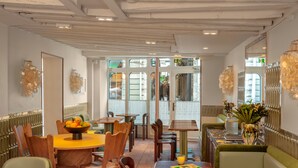 Een restaurantinterieur met houten tafels en stoelen, een ronde tafel in het midden met een fruitschaal, en een groene bank.