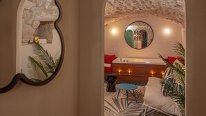 Una habitación acogedora con pared de piedra, un espejo redondo, una cama con almohadones rojos, una mesa redonda azul y una silla blanca.