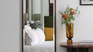 Een slaapkamer met een grote spiegel, een nachtkastje met een bloemenvaas, en een bed met witte lakens.