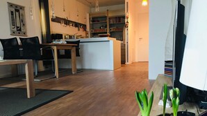 A modern living space with a wooden floor, a dining area with a table and chairs, a kitchen with a white countertop, and a television on the right. - Boardinghouse Oelde - Apartment mit Terrasse (Oelde)